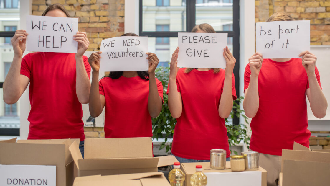 Mindset shifts to unlock your fundraising potential. Charity fundraisers hold signs in front of their faces reading we can help, we need volunteers, please give and be part of it.